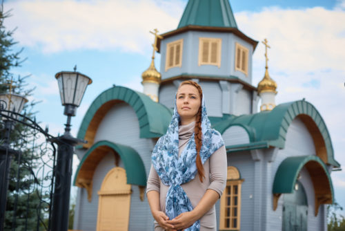 A Russian woman stands in front of a small wooden church. A Russian woman stands in front of a small wooden church.