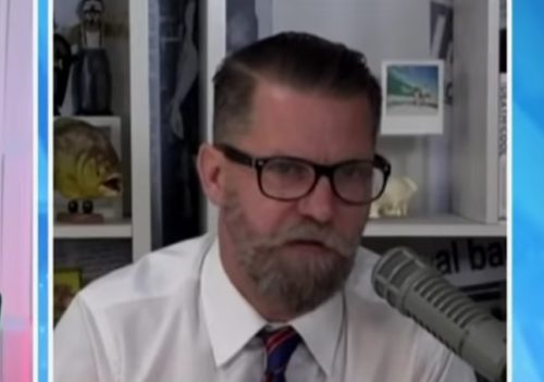 Gavin McInnes in a white shirt and a red tie, talking into a microphone