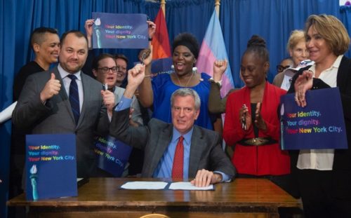Bill de Blasio signed the new rule that allows a third gender option on birth certificates Bill de Blasio sitting, surrounded by people who are cheering.