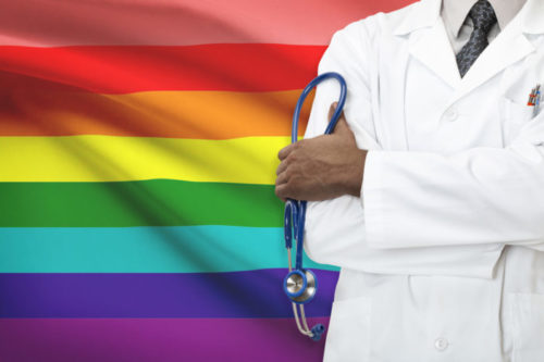 A doctor in front of the 8-striped original version of the rainbow flag.