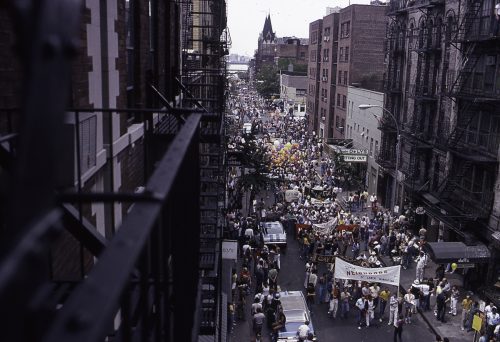 View, looking west, along on Christopher Street (between Hudson and Bleeker streets) of pedestrians, onlookers, and marchers, some with signs and balloons, during the annual New York City Pride March, New York, New York, June 24, 1979. Visible in the distance is the Hudson River.