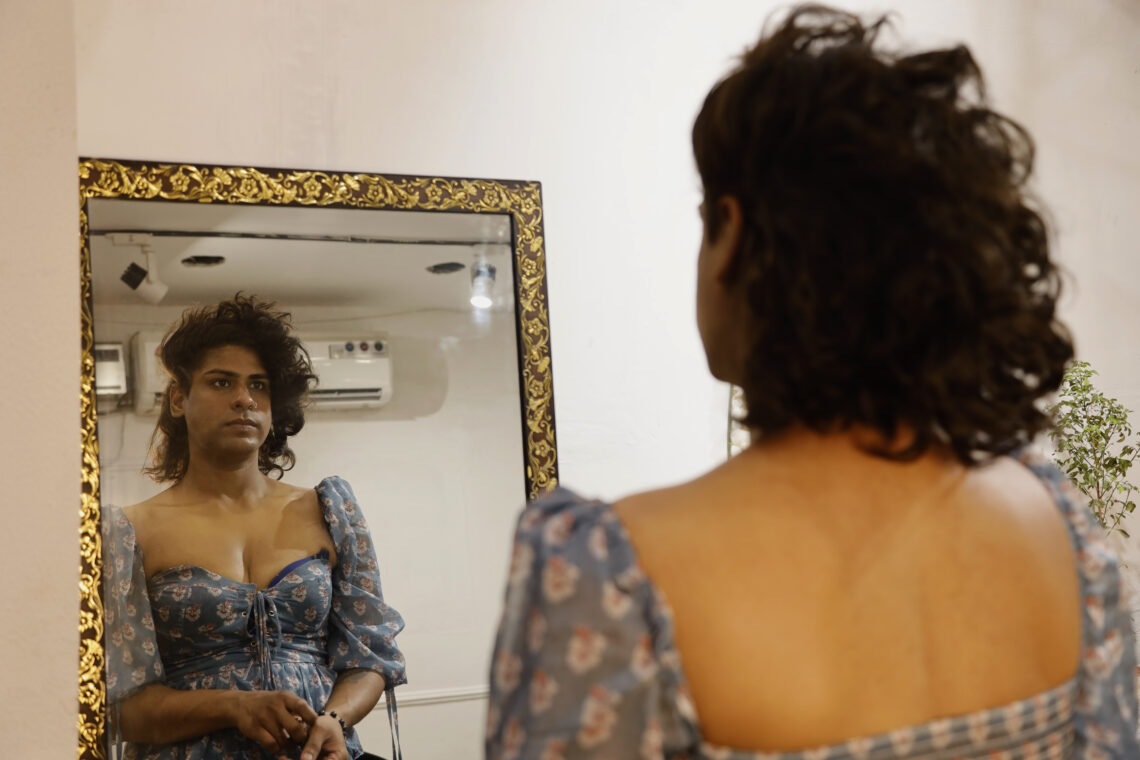 Bruna looks at herself in the mirror as she rubs off her makeup and takes off her wig. She is changing into Akshay for the night.